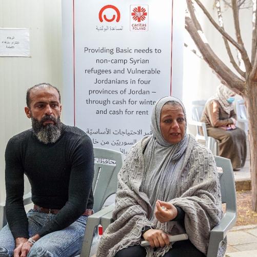 Mężczyzna z zarostem w czarnej bluzce i kobieta z głową i ramionami okrytymi chustami siedzą na krzesłach. W tle znajduje się plakat z logo Polskiej pomocy i Caritas