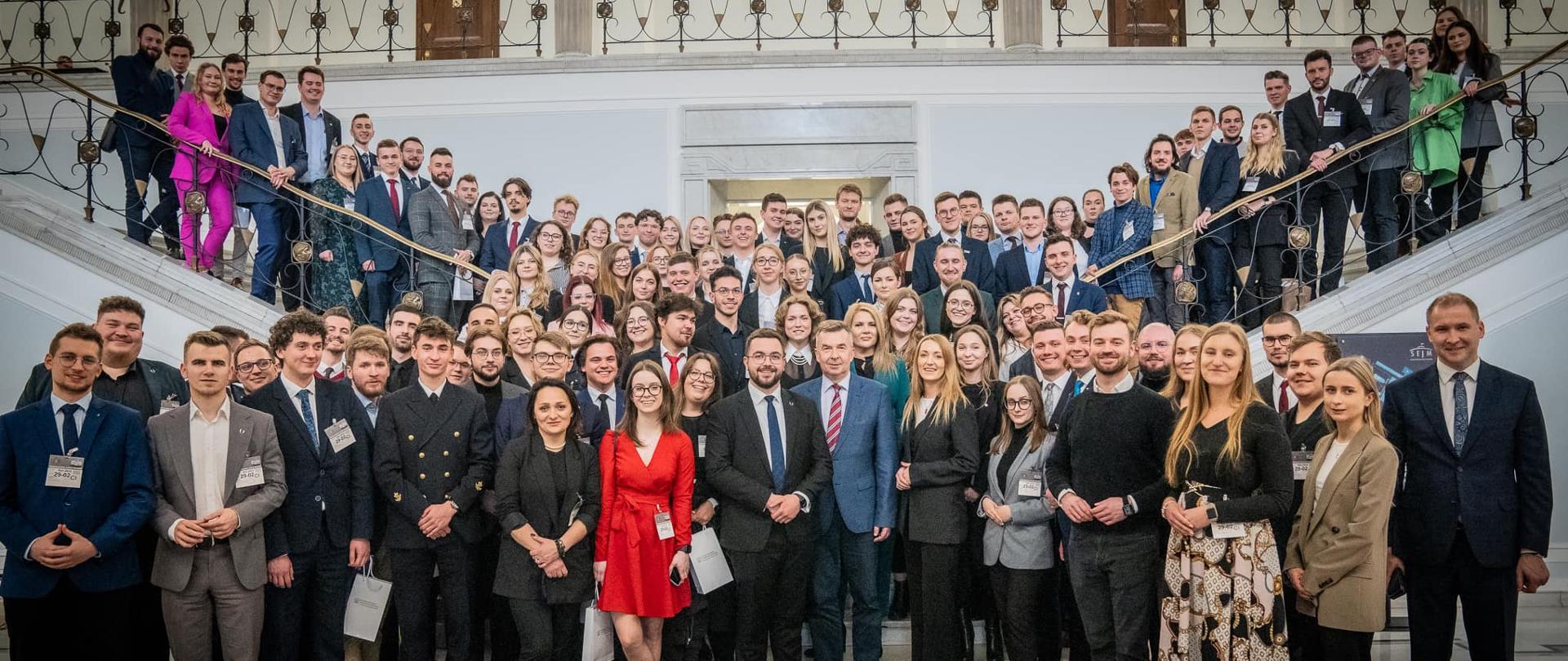 Zdjęcie zbiorowe, na tle schodów stoi duża grupa osób, pośrodku minister Wieczorek, mężczyzna w czarnym garniturze i kobieta w czerwonej sukience.