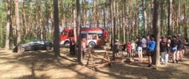 Zdjęcie w lesie. Strażak stoi pośród uczestników obozu harcerskiego i prezentuje wyposażenie strażaków. 