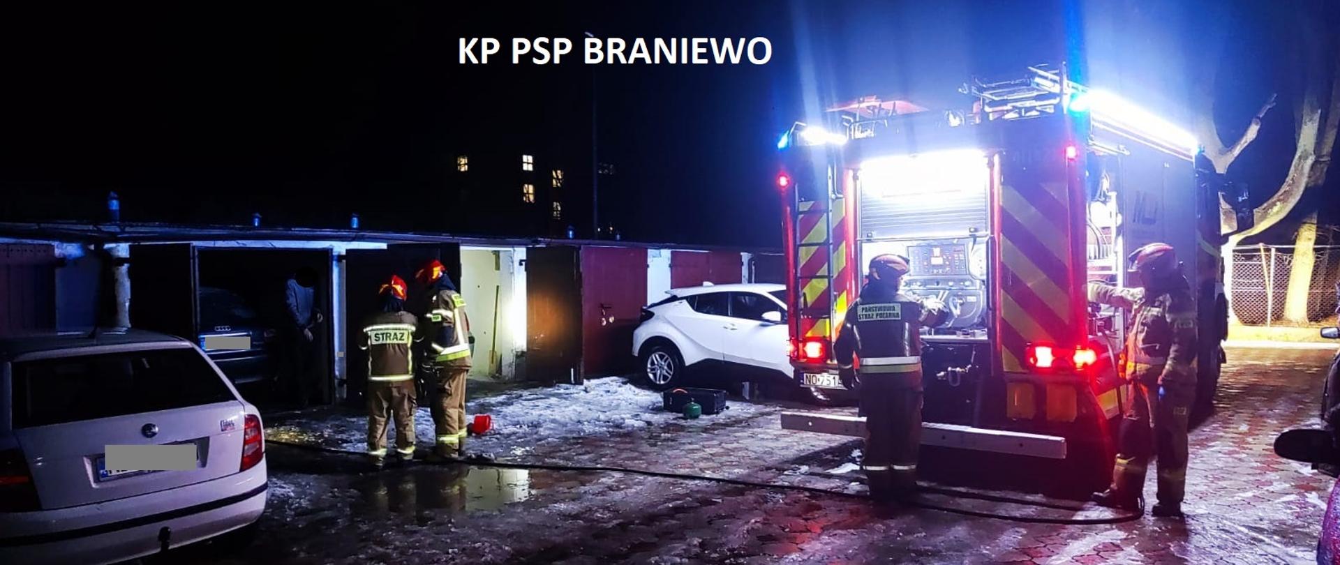 Zdjęcie zrobione nocą. Na zdjęciu stoi stoi samochód strażacki oświetla teren przy garażach. Przy garażach stoją dwa samochody osobowe. Przy samochodzie i garażach strażacy.