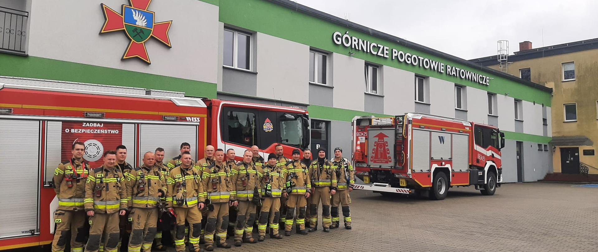 Pamiątkowe zdjęcie strażaków na wybrukowanym placu. Na elewacji budynku widoczny jest duży, zielono-czerwony emblemat ratownictwa górniczego oraz napis: "GÓRNICZE POGOTOWIE RATOWNICZE". Ratownicy w beżowych strojach pozują obok nowoczesnych wozów strażackich marki WISS.