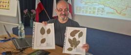 Uśmiechnięty mężczyzna siedzi przy stole i prezentuje dwa arkusze z zasuszonymi liśćmi, umieszczonymi w przezroczystych koszulkach. W lewej ręce trzyma planszę z trzema pojedynczymi liśćmi o różnych kształtach, a w prawej planszę zawierającą większą kolekcję liści. Na stole przed nim stoją laptop, mysz komputerowa oraz butelka wody. W tle znajdują się biało-czerwone flagi i ekran projekcyjny z wyświetloną mapą oraz tabelami. Po lewej stronie kadru widoczna jest tablica z ilustracjami różnych odmian jabłek. Zdjęcie dokumentuje część szkolenia poświęconą identyfikacji odmian drzew owocowych na podstawie cech liści.