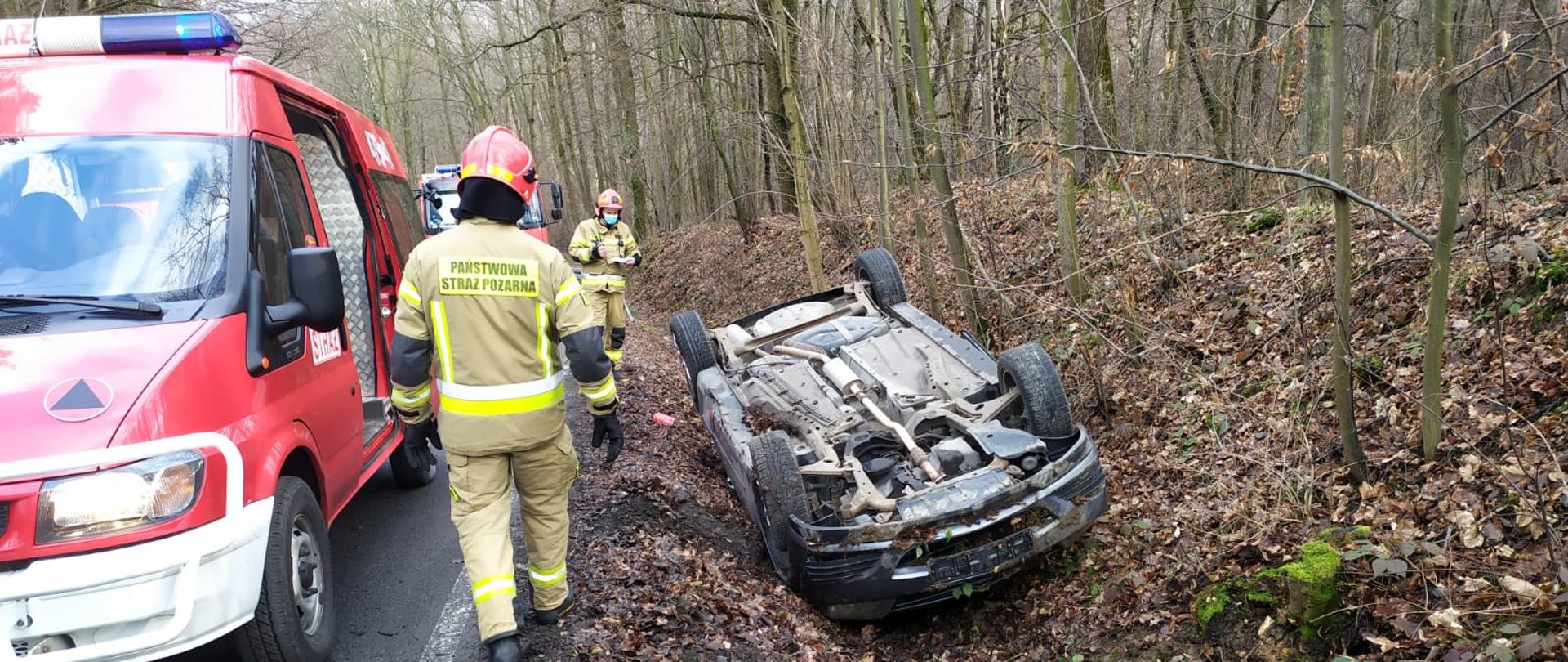 Na zdjęciu widoczny jest samochód osobowy leżący na dachu w przydrożnym rowie. Z lewej strony zdjęcia widoczny jest samochód strażacki oraz strażacy w umundurowaniu specjalnym. Zdarzenie ma miejsce na drodze przebiegającej przez kompleks leśny.