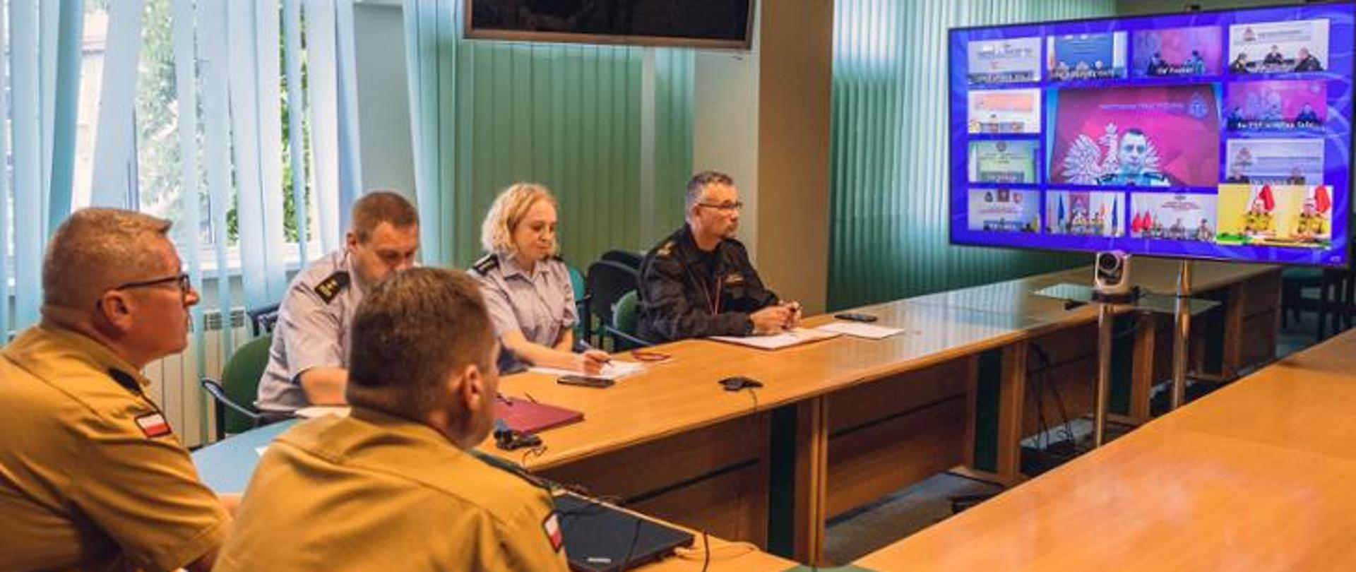 Zdjęcie przedstawia funkcjonariuszy PSP podczas wideokonferencji. Funkcjonariusze siedzą przy stole przed telewizorem na którym widać innych uczestników spotkania.