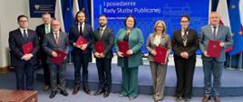 Członkowie nowo powołanej Rady Służby Publicznej w towarzystwie min. Jakuba Stefaniaka, Zastępcy Szefa KPRM oraz min. Anity Noskowskiej-Piątkowskiej, Szefowej Służby Cywilnej - grupa dziewięciu osób stoi przodem do kamery, członkowie RSP trzymają akty powołania w ciemnoczerwonych okładkach, w tle flagi Polski i Unii Europejskiej oraz napis "I posiedzenie Rady Służby Publicznej"