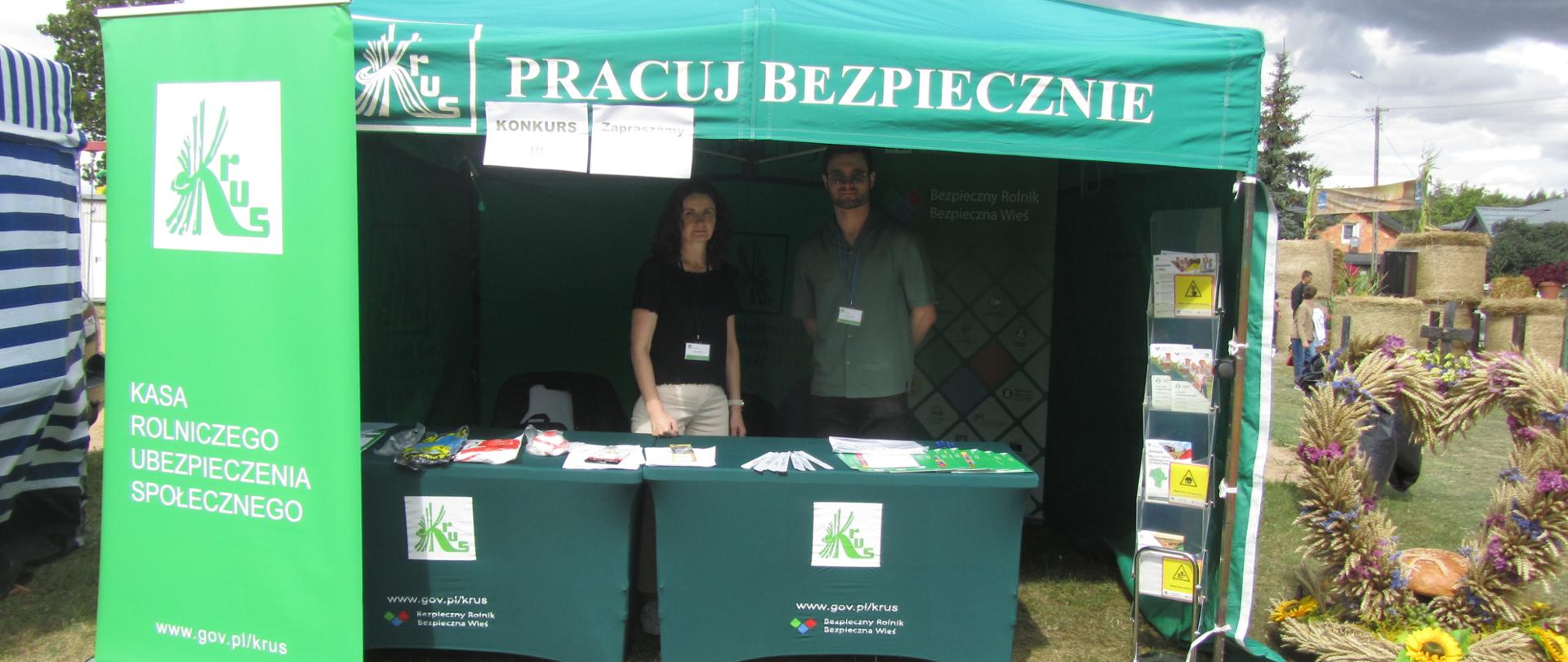 Stoisko KRUS. W centrum zielony namiot opatrzony hasłem: „Pracuj Bezpiecznie”. Po lewej baner z logiem Kasy. Po prawej wieniec dożynkowy. Pod namiotem za stolikami dwoje pracowników KRUS.