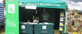 Stoisko KRUS. W centrum zielony namiot opatrzony hasłem: „Pracuj Bezpiecznie”. Po lewej baner z logiem Kasy. Po prawej wieniec dożynkowy. Pod namiotem za stolikami dwoje pracowników KRUS.