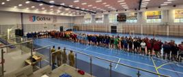 Zdjęcie przedstawia strażaków stojących w hali sportowej podczas otwarcia mistrzostw Wielkopolski Strażaków w Piłce Siatkowej. Zastępca komendanta wojewódzkiego Państwowej Straży Pożarnej otwiera turniej. 