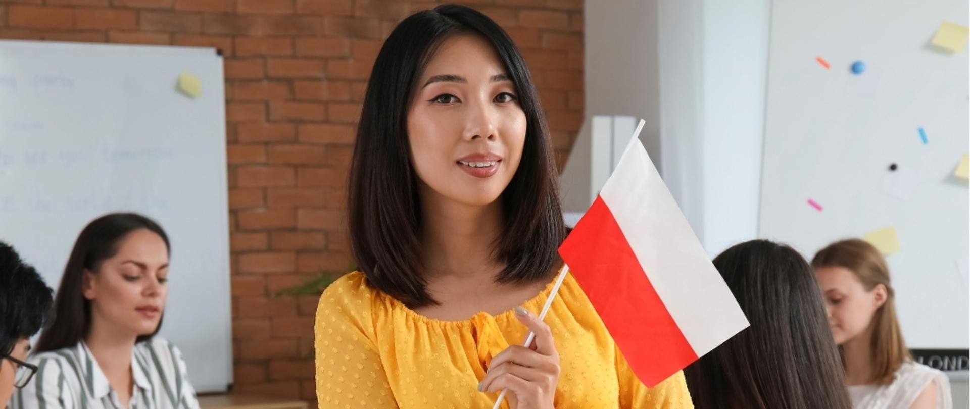 Po środku kobieta w żółtej sukience trzymająca w ręce flagę Polski. W tle widać studentów.