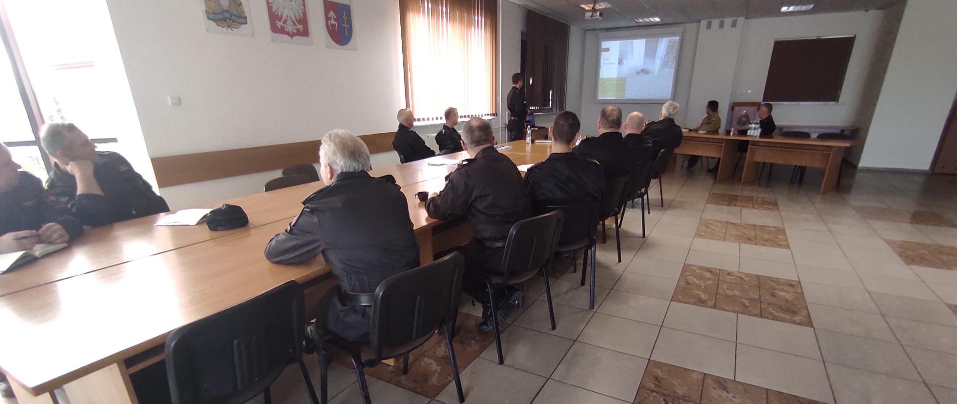 Na zdjęciu strażaków siedzących przy stołach podsumowujących ćwiczenia powiatowe „Mesko 2021”. Jeden z nich przedstawia prezentację z tych ćwiczeń oraz najważniejsze elementy, które byłby poruszane w późniejszej dyskusji.