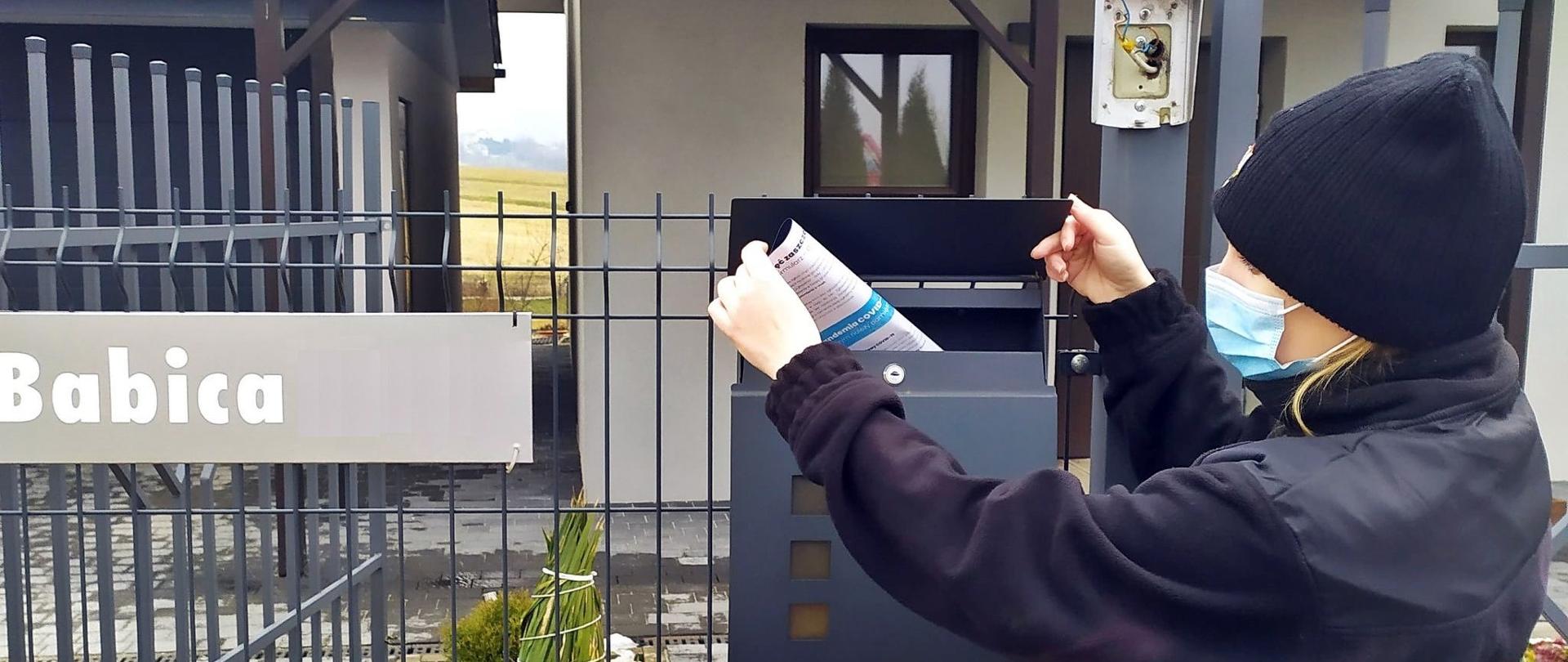 Druhna Ochotniczej Straży Pożarnej w Wysokiej w ubraniu koszarowym stoi przed domem jednorodzinnym i wrzuca ulotkę informacyjną do skrzynki pocztowej umieszczonej na ogrodzeniu budynku mieszkalnego. 