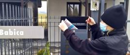 Druhna Ochotniczej Straży Pożarnej w Wysokiej w ubraniu koszarowym stoi przed domem jednorodzinnym i wrzuca ulotkę informacyjną do skrzynki pocztowej umieszczonej na ogrodzeniu budynku mieszkalnego. 