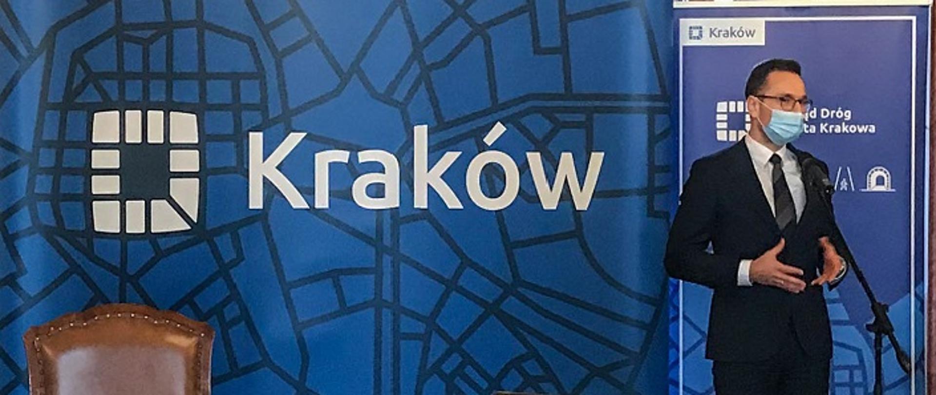 Minister Waldemar Buda stoi przed mikrofonem, na tle ścianki z napisem Kraków. Na pierwszym planie widoczny laptop z transmisją konferencji