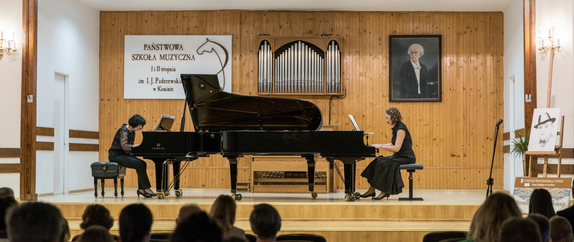Na scenie stoją dwa fortepiany naprzeciw siebie, artystki siedzą w strojach eleganckich.