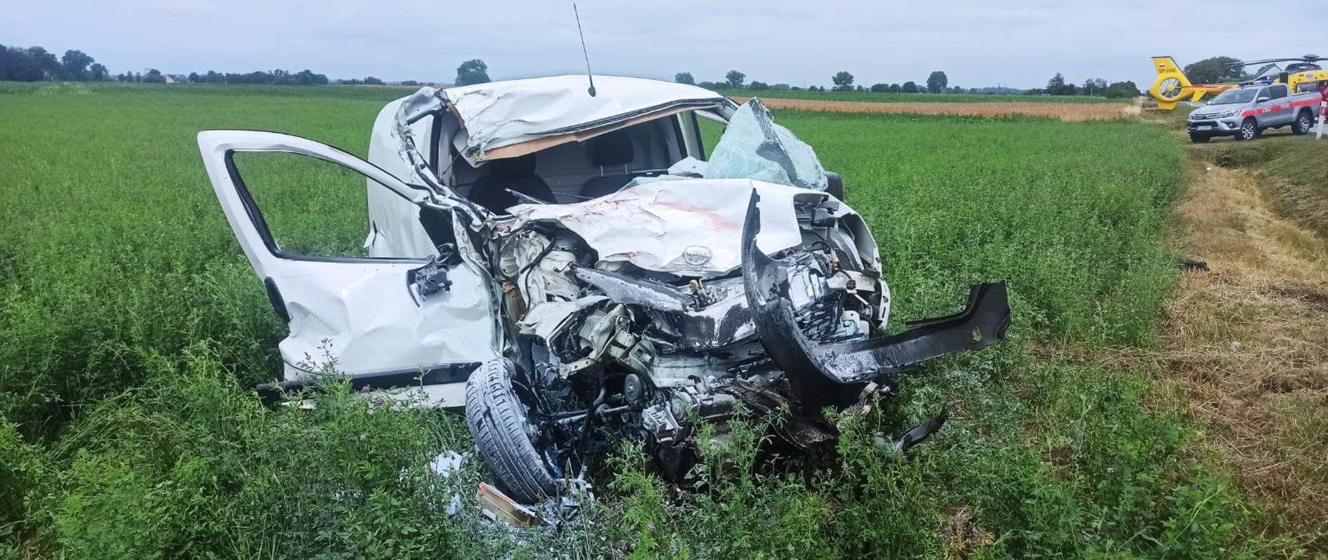 Widać biały uszkodzony samochód od przodu, który znajduje się na polu