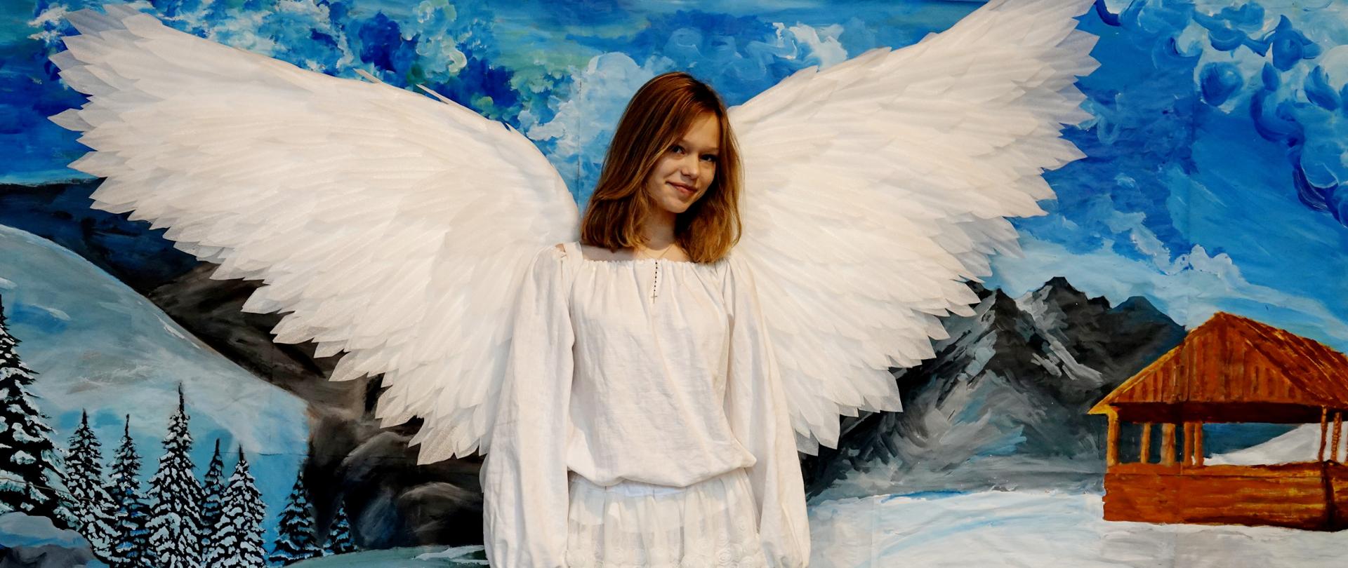 Na zdjęciu uczennica kl. 1A Natalia Putaj. Natalia brała udział w szkolnych Jasełkach. Występuje jako anioł. Dziewczyna ubrana jest w białą bluzkę i białą spódnice. Ma rozpuszczone włosy blond. Dziewczyna ma założone duże białe skrzydła. W tle znajduje się duży obraz na którym namalowany jest pejzaż górski z choinkami, szczytami gór i błękitnym niebem. 