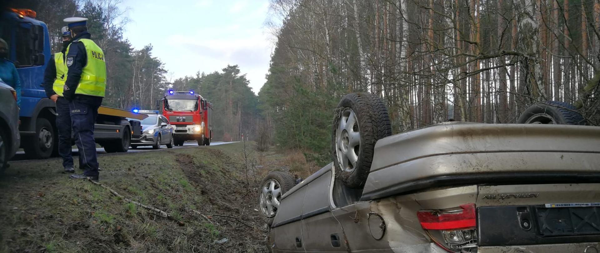 Zdjęcie przedstawia kolizję samochodu osobowego. Na pierwszym planie widać pojazd osobowy na dachu, który uległ kolizji. Na drugim planie widać funkcjonariuszy policji oraz służby ratunkowe po przybyciu na miejsce zdarzenia. Zdjęcie wykonane na poboczu drogi w porze dziennej.