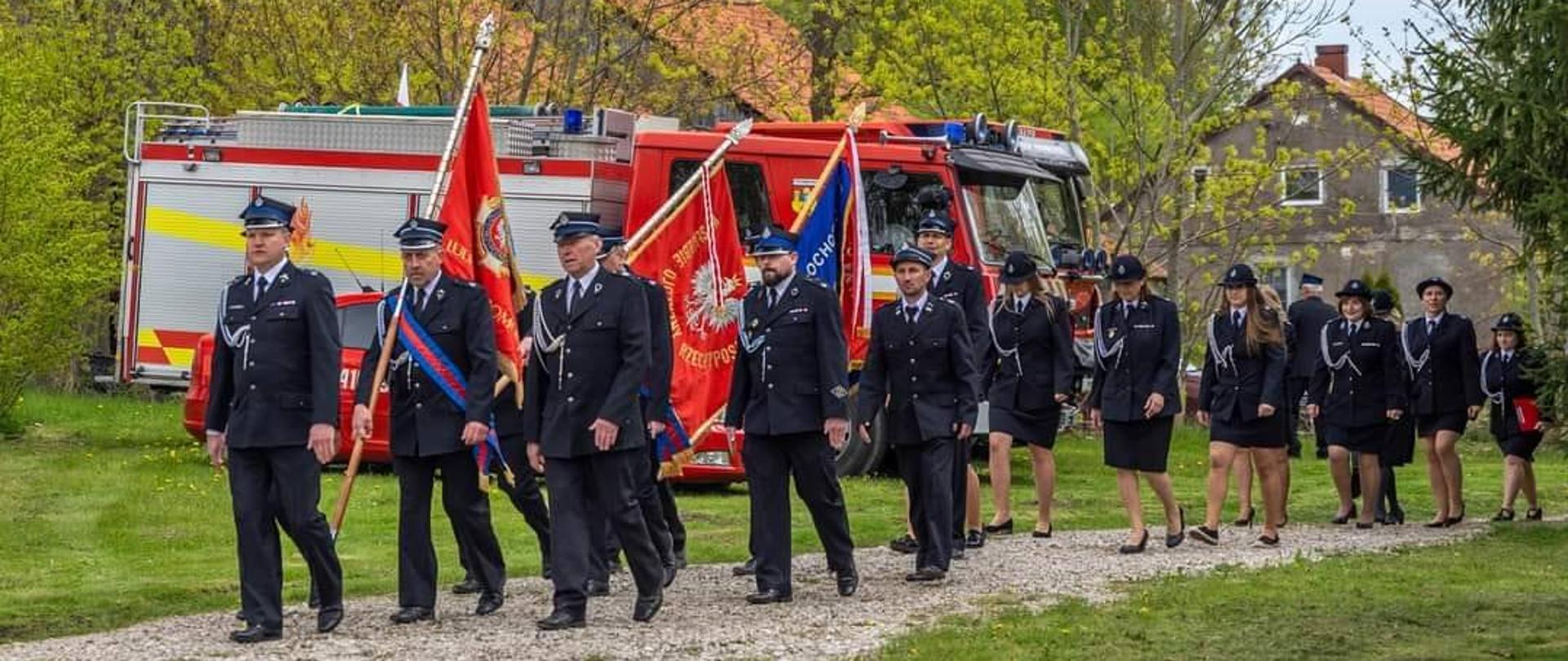 Strażacy ze sztandarami. W tle samochód strażacki.