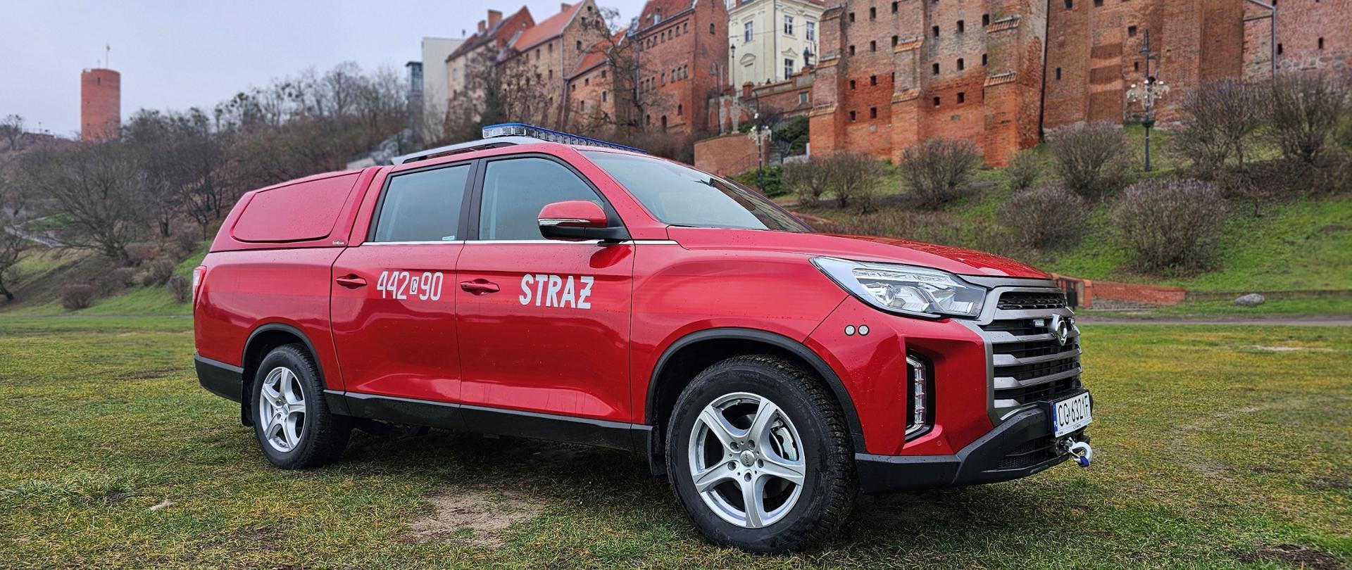 Czerwony strażacki pojazd typu pickup widziany z prawej strony, stojący na trawiastym terenie. W tle gotycka zabudowa starego miasta.