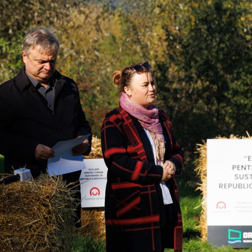 Zdjęcie przedstawia dwie osoby podczas uroczystości. Kobieta przemawia, mężczyzna sprawdza coś na kartce. W tle tablica informacyjna z logo polskiej pomocy 