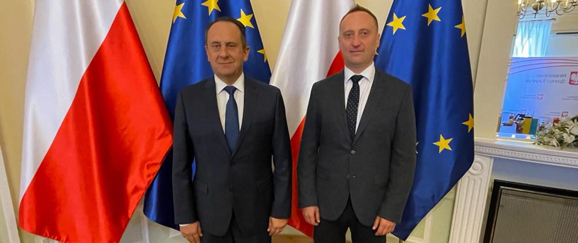 Na zdjęciu znajdują się dwaj elegancko ubrani mężczyźni. Pozują oni na tle czterech flag ustawionych naprzemiennie biało-czerwonej i granatowej z żółtymi gwiazdkami.