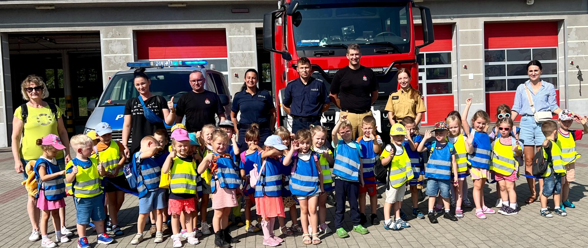 Strażacy, policjanci oraz grupa przedszkolna pozują do zdjęcia na tle komendy oraz samochodu pożarniczego i policyjnego.