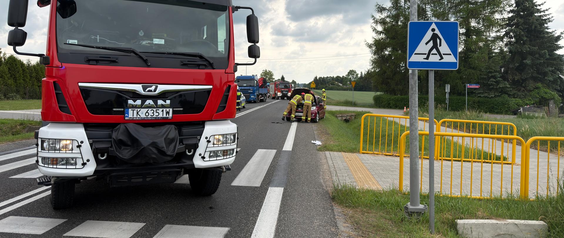Na zdjęciu widać fragment drogi. Widać auto po wypadku oraz oraz dwa wozy strażackie i samochód ciężarowy. Widać strażaków zabezpieczających auto osobowe. 