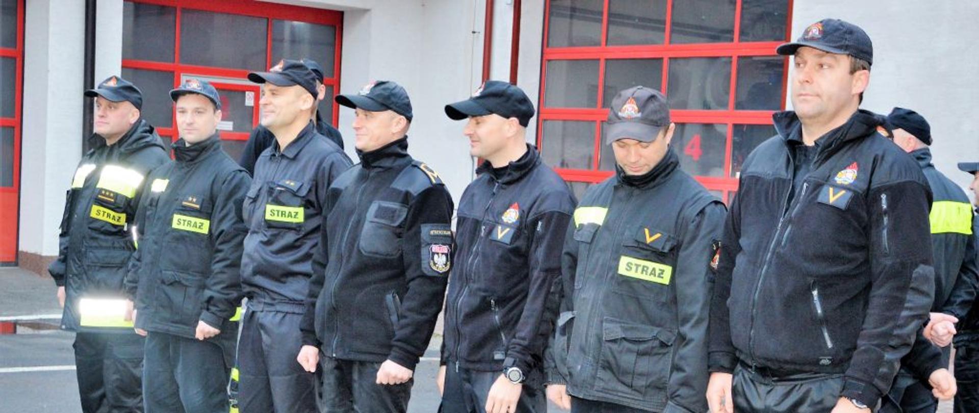 Na zdjęciu widać strażaków którzy zostali awansowani i wystąpili przed szereg w celu odebrania decyzji o awansie. W tle garaże JRG. 