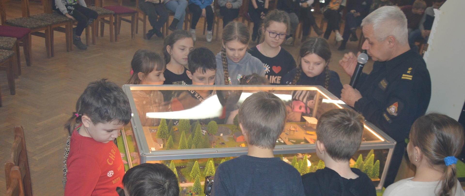 Bezpieczne ferie. Sala wiejska w Sobiałkowie. Wokół symulatora zagrożeń (jezioro-las) znajdują się uczestnicy zajęć - dziewczynki i chłopcy. Strażak - zastępca komendanta powiatowego w ubraniu dowódczo-sztabowym i z mikrofonem w ręce opowiada o zagrożeniach z jakimi można mieć do czynienia podczas wypoczynku w lesie oraz nad jeziorem. W tle pozostali uczestnicy.