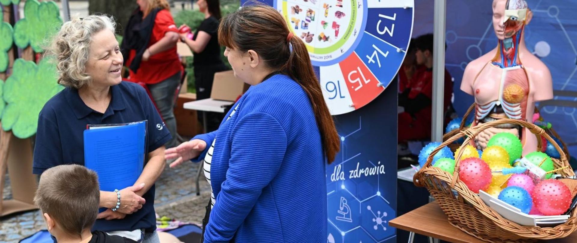 Dwie kobiety rozmawiają przy stoisku edukacyjnym dotyczącym zdrowia; obok widoczny jest model anatomiczny człowieka oraz kosz z kolorowymi piłkami sensorycznymi.

