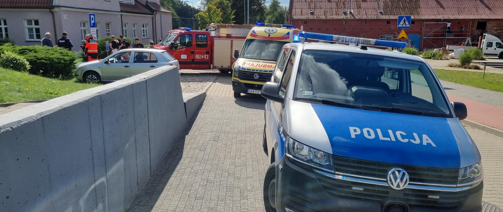 Na chodniku stoi radiowóz, karetka pogotowia oraz samochód ratowniczo-gaśniczy straży pożarnej