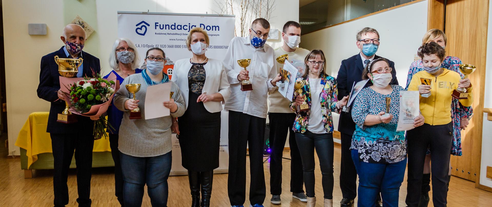 Na zdjęciu laureaci i organizatorzy konkursu plastycznego PFRON, dedykowanego dla osób niepełnosprawnych. Na zdjęciu 11 osób, ustawionych do zdjęcia. W centralnej części kadru wicewojewoda opolski, ubrana w czarną sukienkę i biały żakiet. Po oby stronach stoją laureaci - osoby niepełnosprawne, trzymające w dłoniach puchary i dyplomy pamiątkowe. 