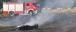 Palące się ściernisko i balot słomy. W tle widać palącą się maszynę rolniczą - balociarkę. Strażacy gaszą pożar. W tle samochód strażacki.