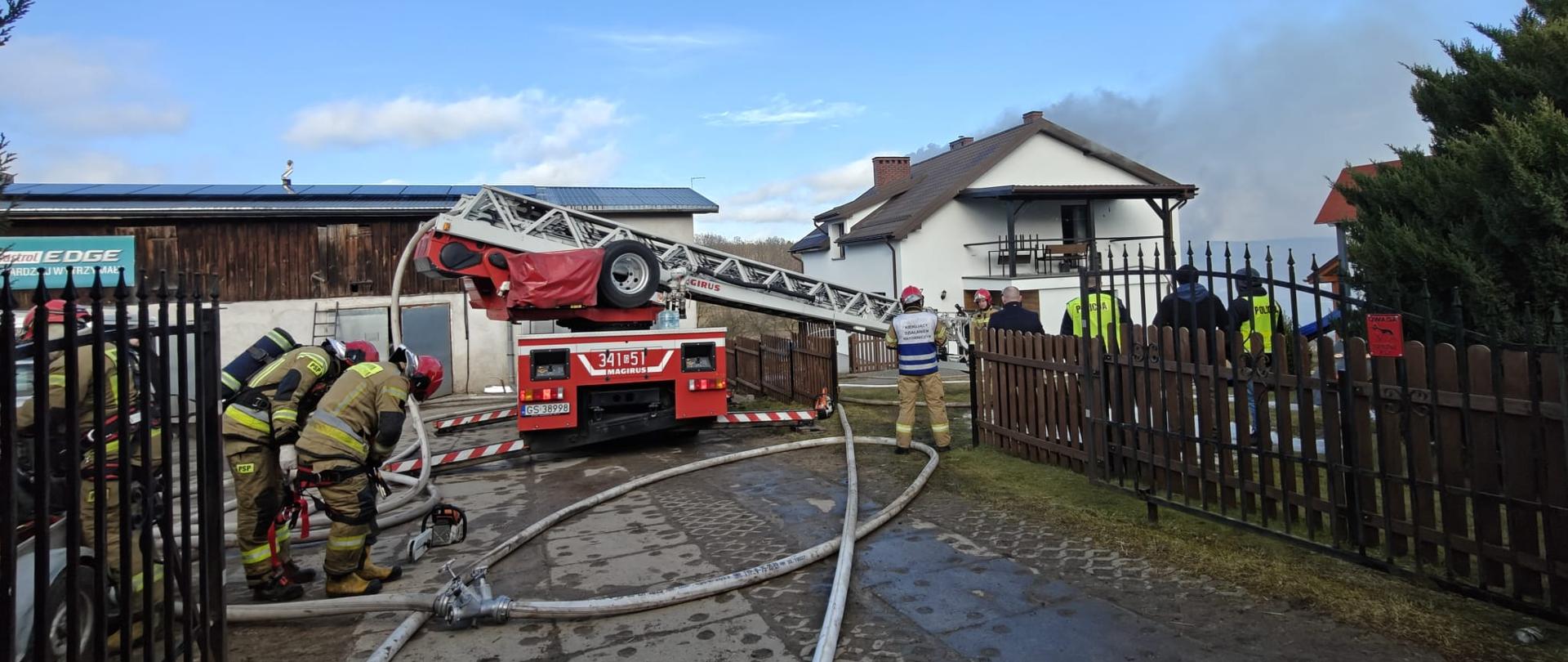 Na zdjęciu strażacy,rozwinięte linie gaśnicze, drabina strażacka mechaniczna, w oddali palący się dom