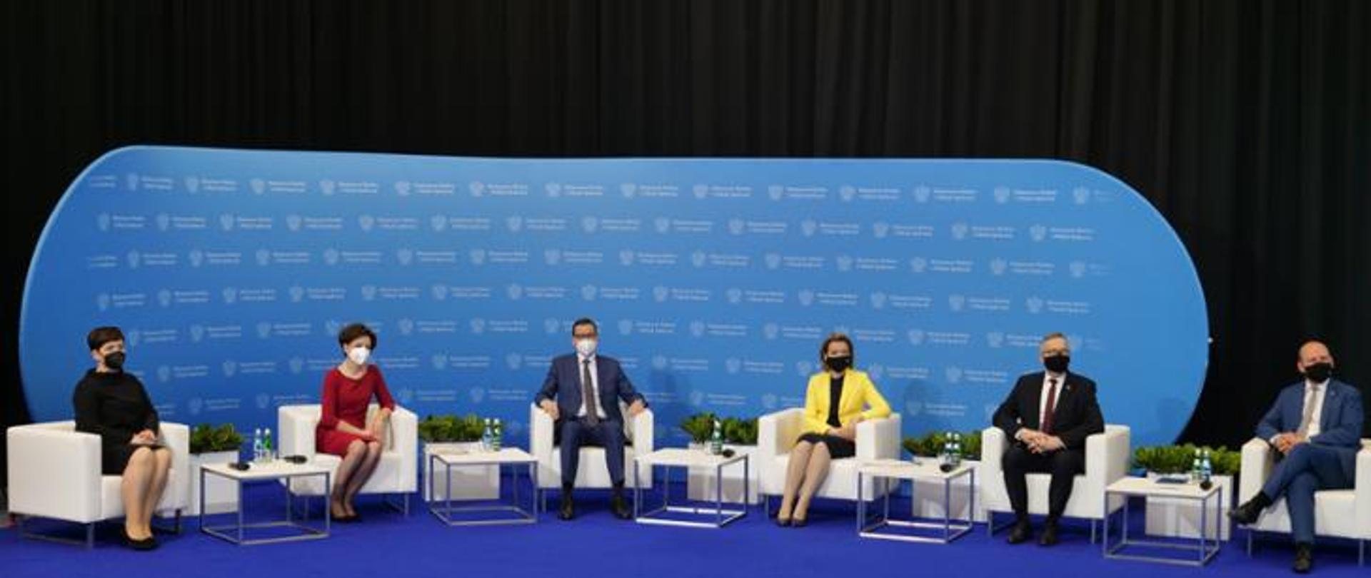 Niebieski dywan. Na nim sześć białych foteli. W fotelach siedzą ludzie. W tle na niebieskim tle słabo widoczne logo Ministerstwa Rodziny i Polityki Społecznej.