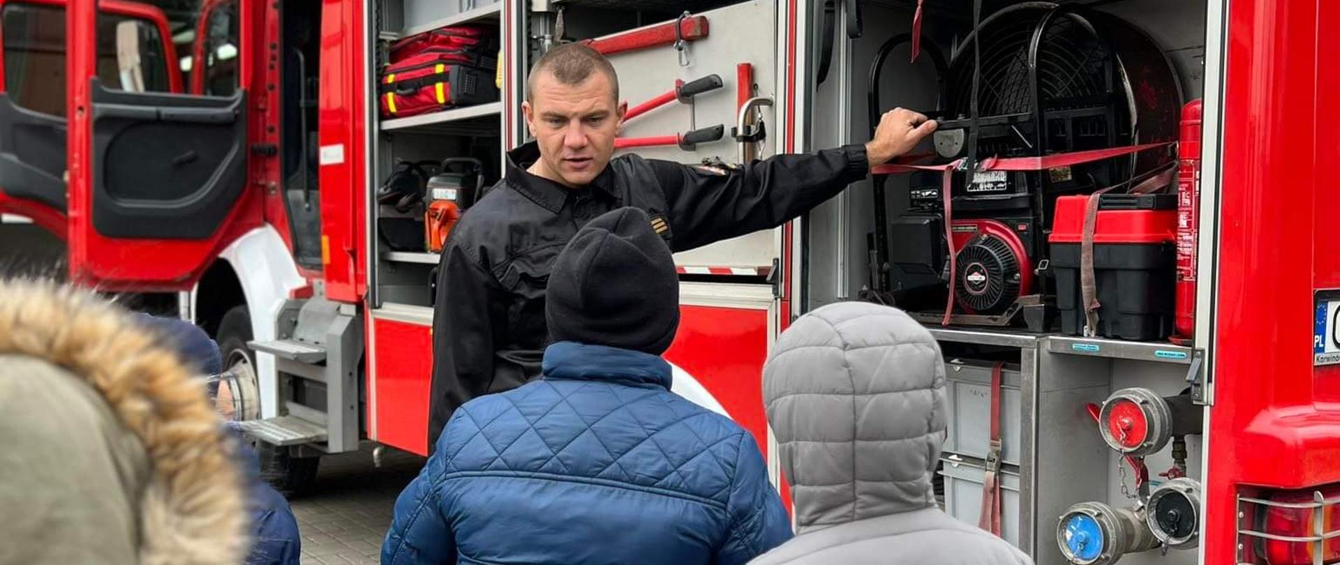 Zdjęcie przedstawia grupę dzieci stojących tyłem .Z przodu strażak w umundurowaniu koszarowym prezentujący dzieciom sprzęt strażacki