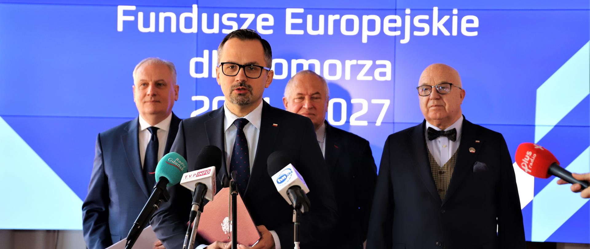 Wiceminister Marcin Horała stoi przed mikrofonem. Za jego plecami stoją trzy osoby.