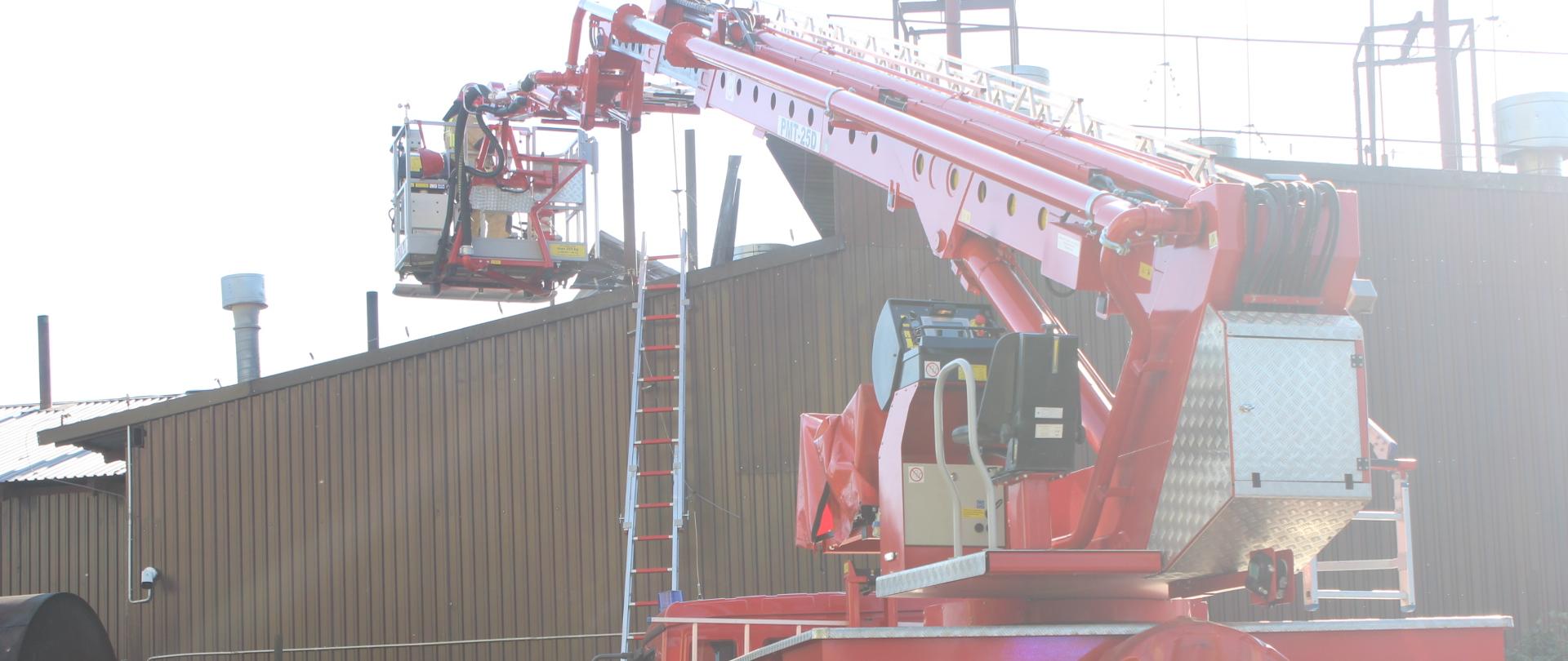 Podnośnik hydrauliczny rozstawiony przed budynkiem, w którym miał miejsce pożar. W koszu podnośnika strażak.