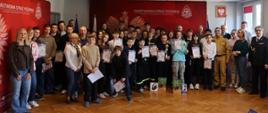 To zdjęcie stanowi zbiorowe podsumowanie wydarzenia, gromadząc wszystkich uczestników i organizatorów na wspólnej fotografii pamiątkowej w siedzibie Państwowej Straży Pożarnej w Dzierżoniowie. Liczna grupa dzieci i młodzieży wypełnia niemal całą szerokość sali, pozując na tle dwóch czerwonych ścianek promocyjnych z godłem państwowym.
Wielu uczniów dumnie prezentuje otrzymane dyplomy, a laureaci pierwszych miejsc trzymają w dłoniach złote puchary. Na pierwszym planie widoczne są również nagrody rzeczowe przygotowane dla zwycięzców. Młodzieży towarzyszą dorośli: opiekunowie w strojach cywilnych oraz funkcjonariusze straży pożarnej w pełnym umundurowaniu, co podkreśla rangę i profesjonalny charakter spotkania.
Całość została uwieczniona w jasnej, przestronnej sali z tradycyjnym parkietem. Zdjęcie doskonale oddaje skalę przedsięwzięcia oraz radosną, a jednocześnie pełną powagi atmosferę towarzyszącą finałowi turnieju.