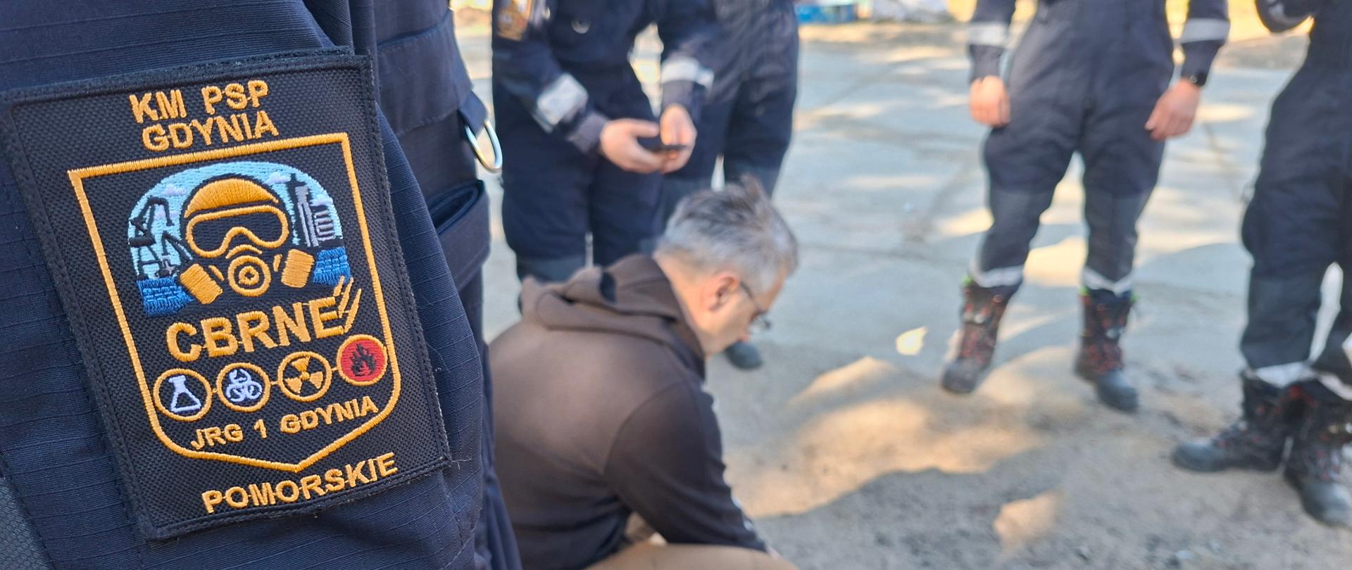 Strażacy stojący w okręgu w ubraniach specjalnych. Jeden z nich pobiera próbkę z ziemi.