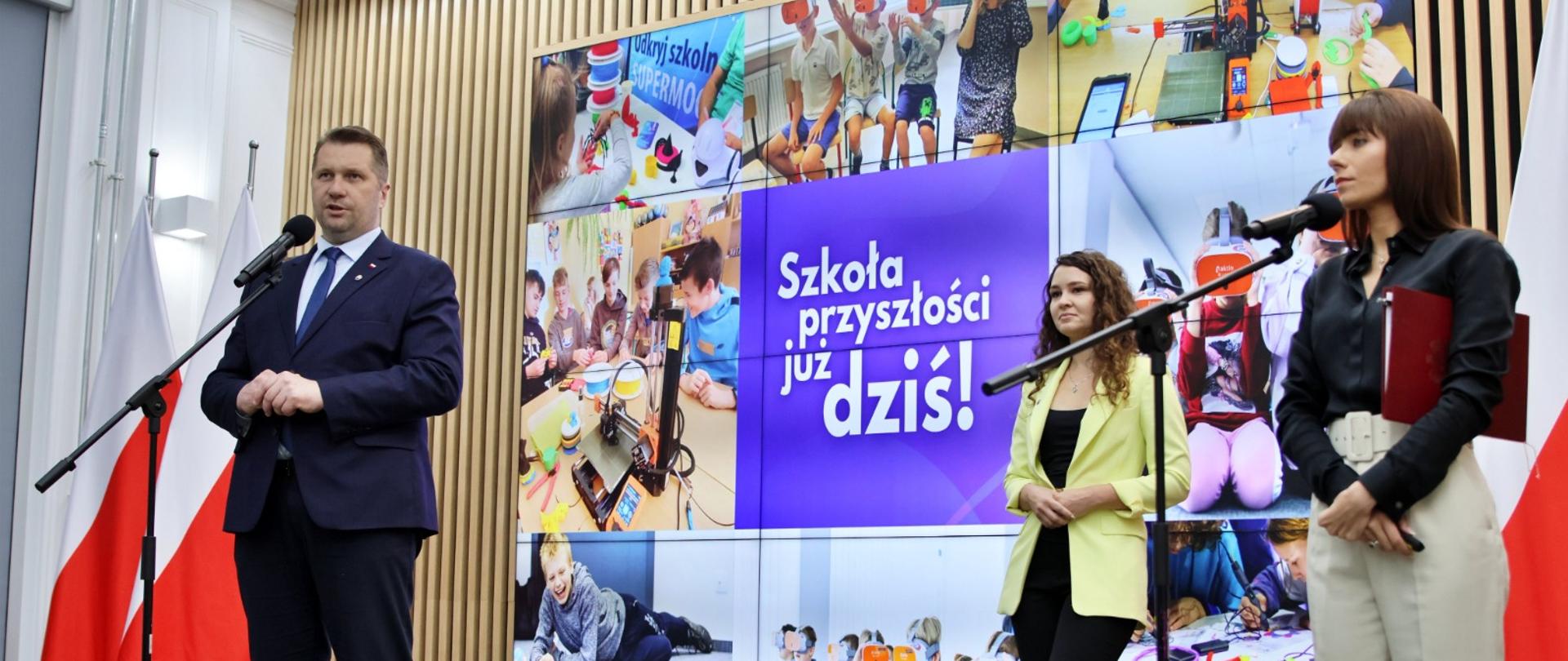 Na sali stoi minister Czarnek i mówi do mikrofonu na stojaku, obok niego dwie kobiety, za nimi wielki ekran z napisem Szkoła przyszłości już dziś.