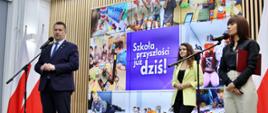 Na sali stoi minister Czarnek i mówi do mikrofonu na stojaku, obok niego dwie kobiety, za nimi wielki ekran z napisem Szkoła przyszłości już dziś.