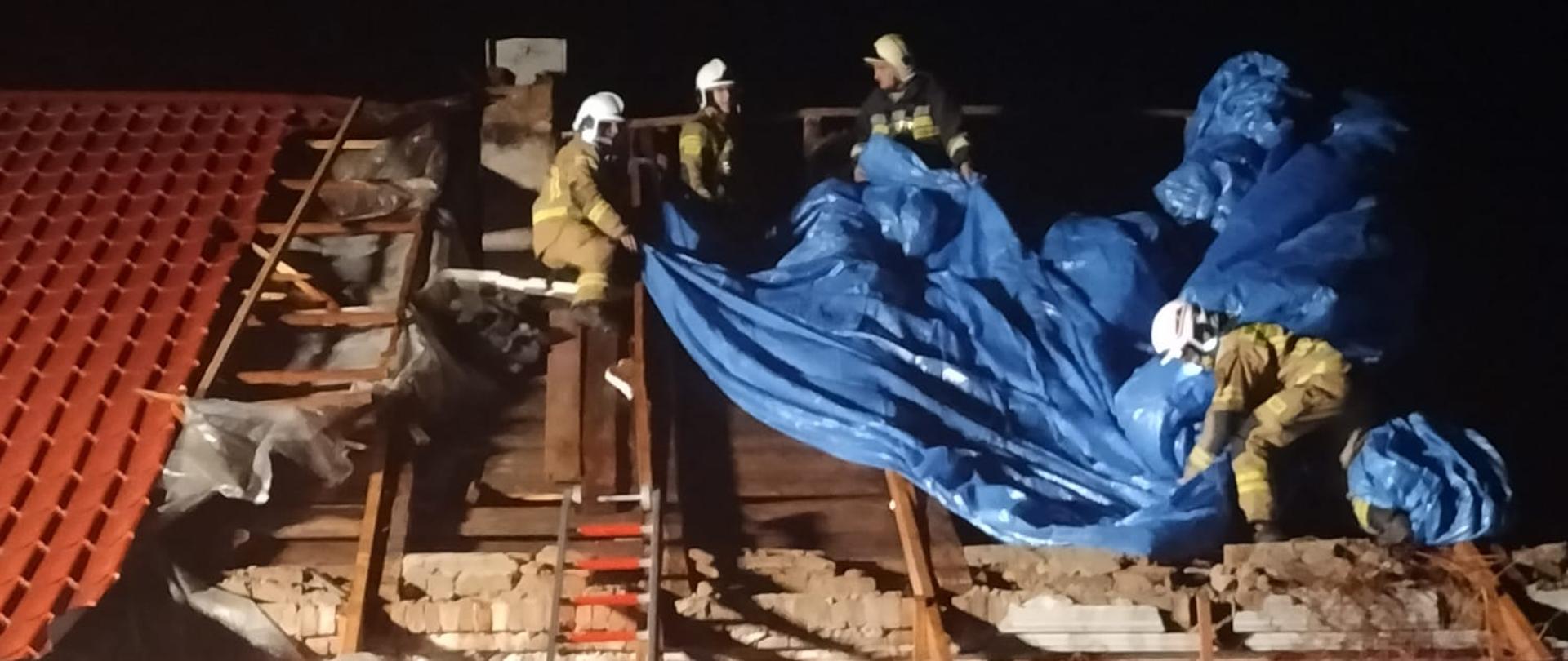 Na zdjęciu widzimy strażaków pracujących przy zabezpieczeniu plandeką dachu budynku mieszkalnego. Dach uszkodzony w wyniku porywistych wiatrów.