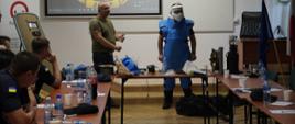 Siedzący w sali wykładowej uczestnicy szkolenia patrzą się na instruktora oraz stojącego obok mężczyznę w kombinezonie i masce
