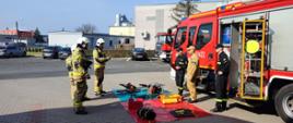 Zdjęcie zrobione na zewnątrz budynku na placu KP PSP w Środzie WLKP. w tle widać zabudowania oraz cukrownię. Na placu stoi samochód pożarniczy z podniesionymi roletami, przed nim znajdują się funkcjonariusze PSP oraz pole sprzętowe z zestawem hydraulicznym oraz pneumatycznym. Naprzeciwko samochodu pożarniczego znajdują się egzaminowani strażacy OSP w ubraniach bojowych.