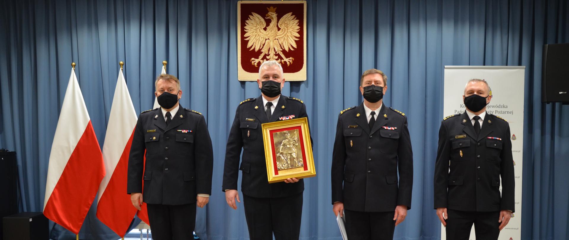 W szeregu stoi czterech strażaków w umundurowaniu galowym, drugi od lewej trzyma pamiątkowy wizerunek św. Floriana oprawiony w ramki w tle widać niebieską zasłonę, trzy flagi państwowe oraz wiszące godło polski.