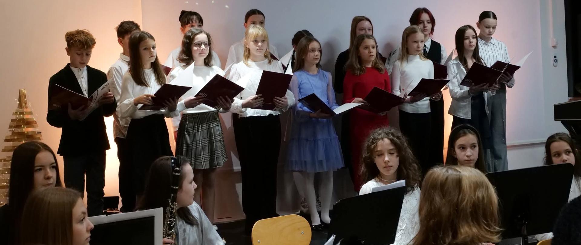 Na zdjęciu widoczna jest scena z muzykami którzy siedzą przy pulpitach a za nimi stoi chór z otwartymi teczkami w rękach w kolorze czarnym. W tle wyświetlana jest na ścianie dekoracja w postaci masek.