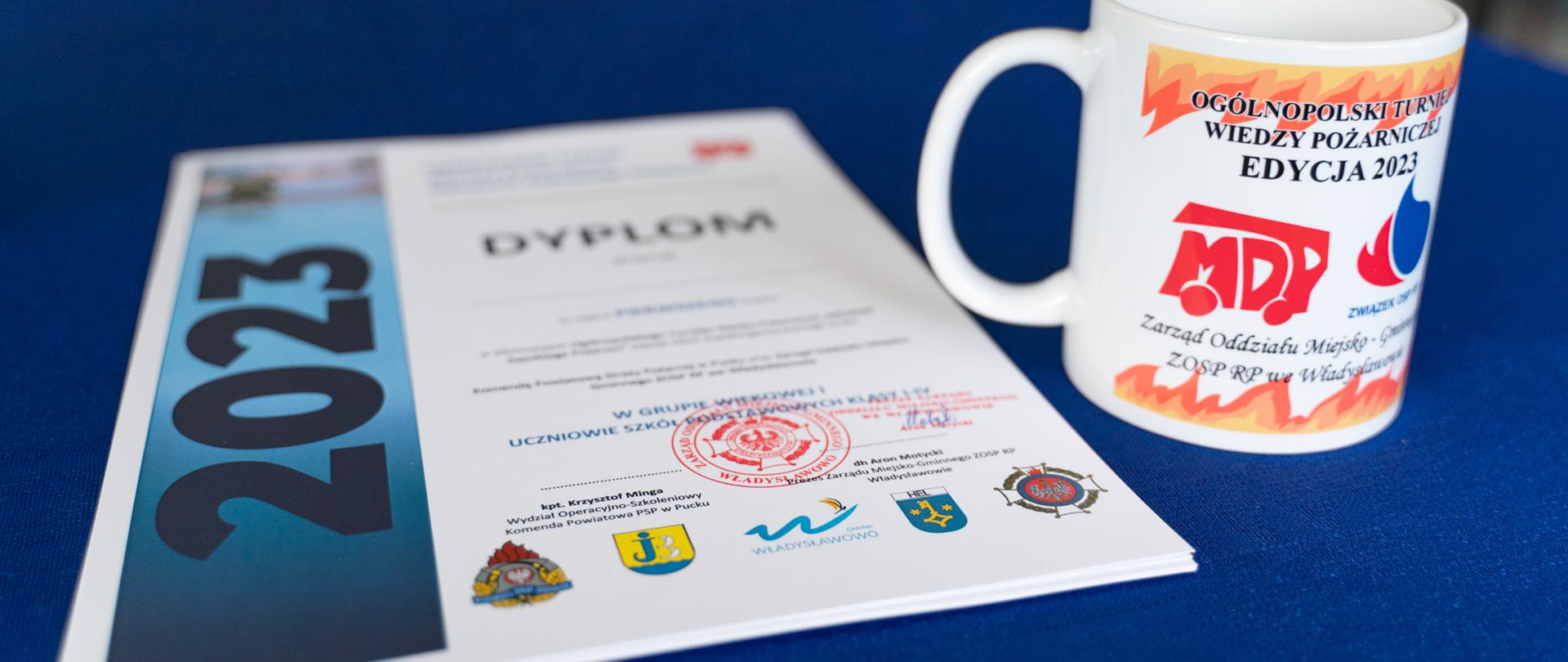 Zdjęcie przedstawia dyplom i kubek z logo OGÓLNOPOLSKIEGO TURNIEJU WIEDZY POŻARNICZEJ