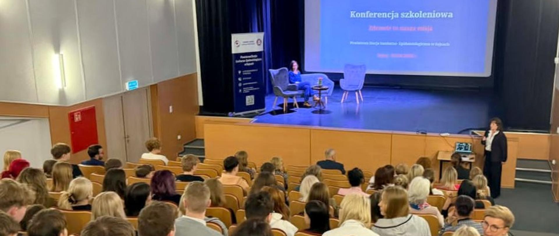 Młodzież siedząca w auli, na scenie osoba siedząca na kanapie, w tle duży ekran z napisem "Konferencja szkoleniowa"