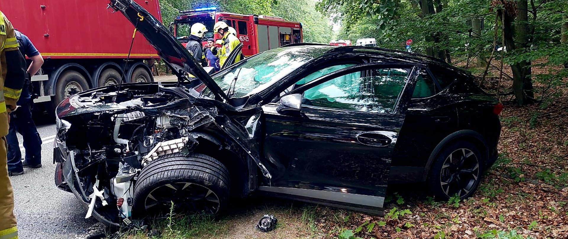 Zdjęcie przedstawia rozbite auto oraz służby działające na miejscu zdarzenia.
